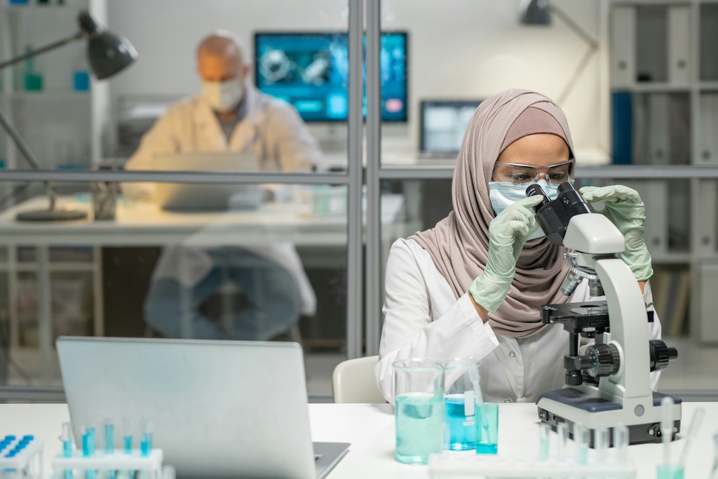 A female scientist looking down a microscope, surrounded by vials of blue liquid